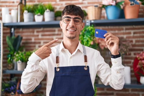 Giovane uomo ispanico che lavora al negozio di fiorista in possesso di carta di credito sorridente felice indicando con mano e dito  - Foto, immagini