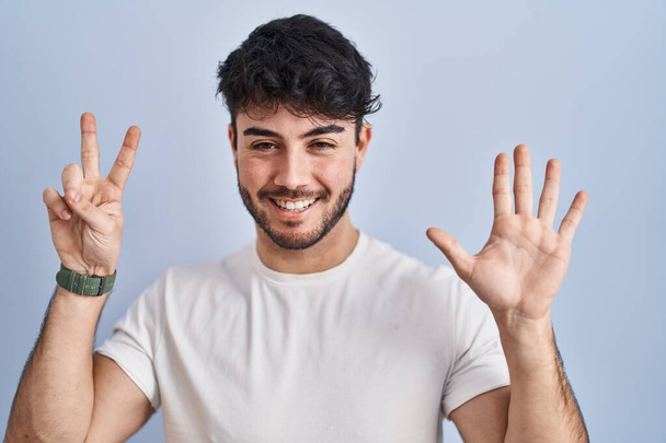 Латиноамериканець з бородою, що стоїть над білим фоном, показує і вказує вгору з пальцем номер сім, посміхаючись упевнено і щасливо.  - Фото, зображення
