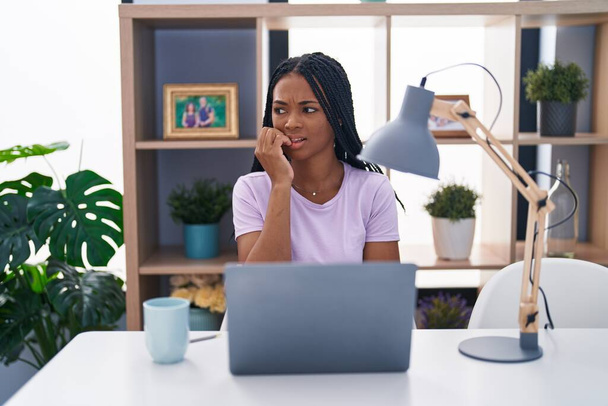 Afroamerykanka z warkoczami używająca laptopa w domu wyglądająca na zestresowaną i zdenerwowaną z rękami obgryzającymi paznokcie. niepokój.  - Zdjęcie, obraz