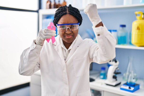 Femme afro-américaine travaillant au laboratoire scientifique tenant un ruban rose agacé et frustré criant de colère, criant fou de colère et la main levée  - Photo, image