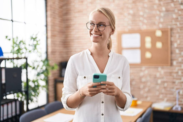 Jonge blonde vrouw zakenman met behulp van smartphone werken op kantoor - Foto, afbeelding