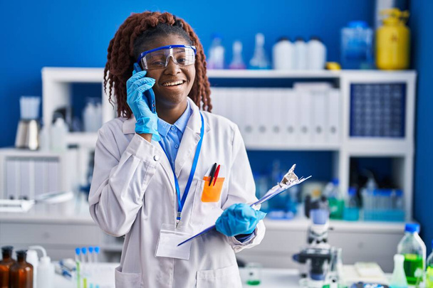 African american woman scientist talking on smartphone reading document at laboratory - Photo, Image