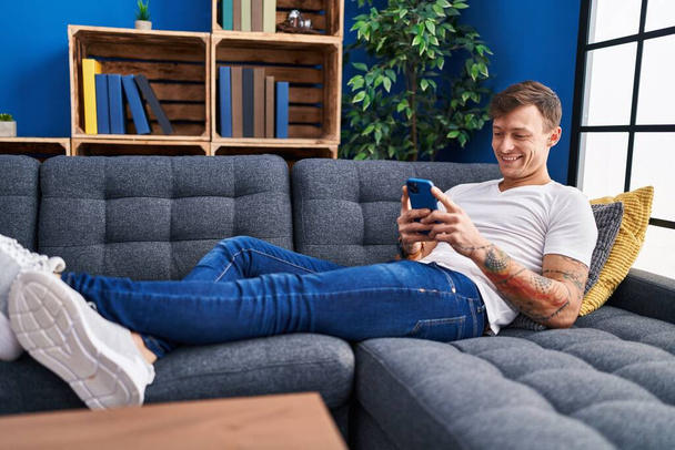 Hombre joven usando teléfono inteligente tumbado en el sofá en casa - Foto, Imagen