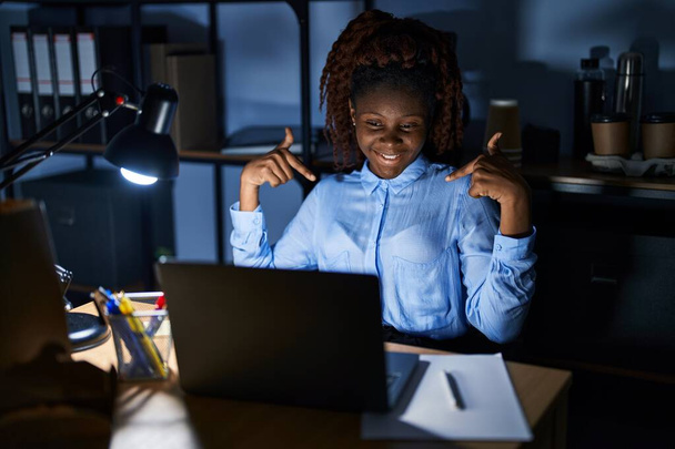 Африканська жінка працює в офісі вночі, дивлячись впевнено з посмішкою на обличчі, вказуючи на себе з пальцями гордими і щасливими.  - Фото, зображення