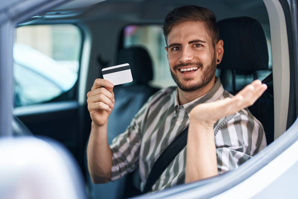 Spanischer Mann mit Bart fährt Auto und hält Kreditkarte und feiert Leistung mit glücklichem Lächeln und Siegerausdruck mit erhobener Hand  - Foto, Bild