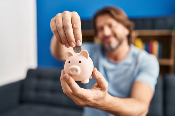 Middle age man inserting coin on piggy bank sitting on sofa at home - Foto, immagini