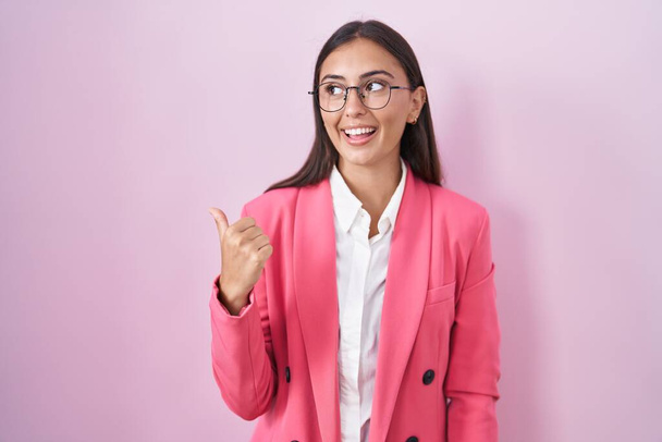 Young hispanic woman wearing business clothes and glasses smiling with happy face looking and pointing to the side with thumb up.  - Foto, Imagen