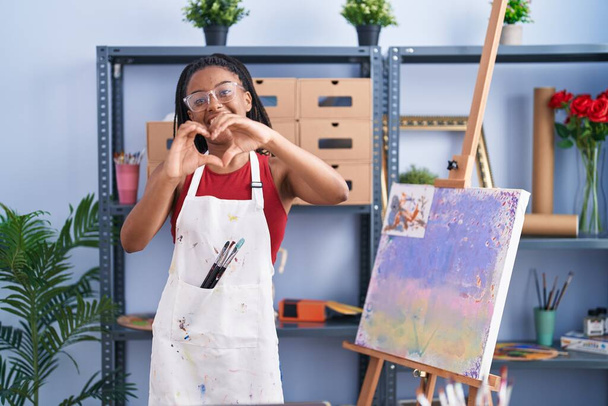 Młody afrykański Amerykanin z warkoczami w pracowni malarstwa artystycznego na płótnie uśmiechający się w miłości pokazując symbol serca i kształt z rękami. romantyczna koncepcja.  - Zdjęcie, obraz