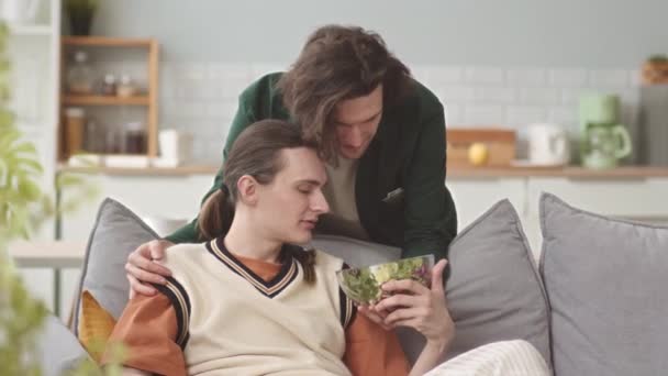 Турботливий молодий красивий гей чоловік приносить чашу свіжого зеленого салату своєму партнерові, відпочиваючи разом у затишній квартирі - Кадри, відео