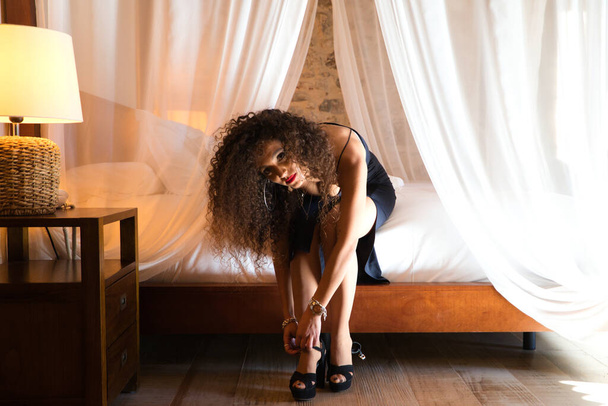 beautiful brunette woman with curly hair sitting on the bed fastening her high heels. The woman is wearing an elegant black dress with red lipstick. - Photo, Image