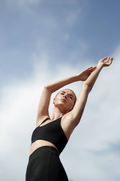 Ένα ξανθό μοντέλο γυμναστικής με μαύρα αθλητικά που απλώνει τα χέρια της προς τον ουρανό. Στέκεται απέναντι σε έναν γαλάζιο ουρανό με άσπρα σύννεφα - Φωτογραφία, εικόνα