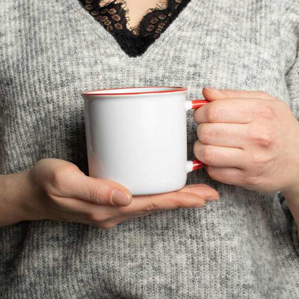 Női kezek közelsége kerámia korsó itallal. Gyönyörű lány kezében egy fehér piros csésze teát vagy kávét a reggeli napfényben. Bögrét a tervedhez. Üres mockup sablon - Fotó, kép
