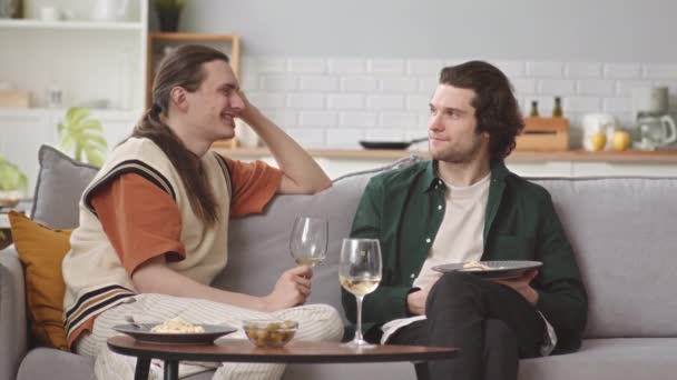 Ρομαντικό ομοφυλόφιλο ζευγάρι κάθεται στον καναπέ στο σαλόνι κουβεντιάζοντας ενώ έχει δείπνο με λευκό κρασί - Πλάνα, βίντεο