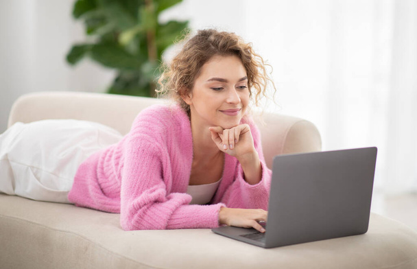 Schöne junge Frau, die es sich auf der Couch mit dem Laptop bequem macht, glückliche Millennial-Frau, die auf dem Sofa im gemütlichen Wohnzimmer zu Hause liegt, im Internet surft, Online-Shopping macht, Kopierraum - Foto, Bild