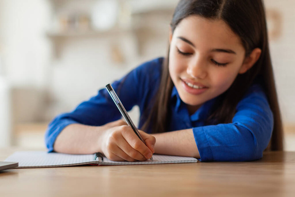 Домашнее задание. Улыбающаяся школьница пишет или рисует в бумажной тетрадке, сидя за столом дома. Девочка делает заметки за столом. Образование и досуг. Селективный фокус - Фото, изображение