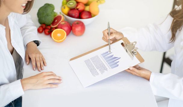 Dospělý bělošský lékař výživy v bílém plášti ukazuje plán hubnutí mladé pacientce v interiéru kliniky. Řádná výživa, doporučení a rady ze strany odborníků, zdravotní péče - Fotografie, Obrázek