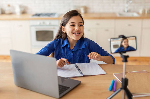 Modern E-Learning. Vrolijk Arabisch schoolmeisje met online les via video bellen op mobiele telefoon, met behulp van laptop en notities maken, Studeren op afstand kijken educatieve lezing thuis - Foto, afbeelding