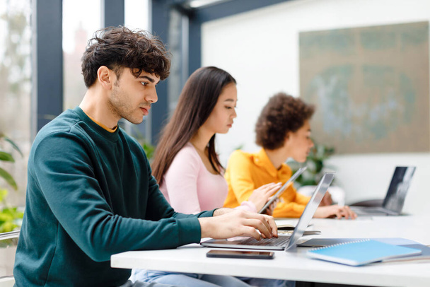 Étudiants multiraciaux masculins et féminins étudiant pour le test, faire des devoirs avec des gadgets, gars concentré à l'aide d'un ordinateur portable, assis à table dans un espace de coworking - Photo, image