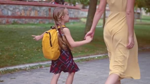 Feliz colegiala sostiene la mano de la madre ir a la escuela juntos en el parque de la ciudad mujer lleva a su hija a la escuela caminando por el jardín urbano relaciones familiares - Imágenes, Vídeo