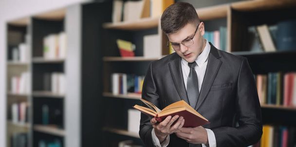 Een mannelijke advocaat of manager leest een leerboek op de achtergrond van een plank met literatuur. - Foto, afbeelding
