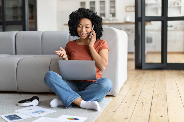 Zdalny biznes. Czarna freelancerka używająca laptopa i mówiąca przez komórkę w domu, uśmiechnięta Afroamerykanka pracująca zdalnie z komputerem i papierami siedząc na podłodze w salonie - Zdjęcie, obraz