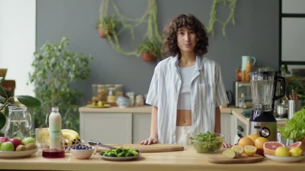 Νεαρή γυναίκα blogger που περπατά στην κουζίνα, χαιρετώντας τους θεατές και μιλώντας για smoothie συστατικά στην κάμερα, ενώ κινηματογραφεί υγιεινή συνταγή βίντεο για τη διατροφή vlog στο σπίτι - Πλάνα, βίντεο