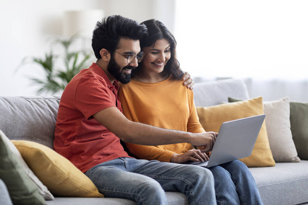 Mosolygó fiatal indiai házastársak segítségével Laptop Számítógép otthon együtt, Boldog Millenniumi keleti pár pihentető kanapén, Online vásárlás vagy foglalás nyaralás, Férfi mutató eszköz képernyő, Szabad hely - Fotó, kép