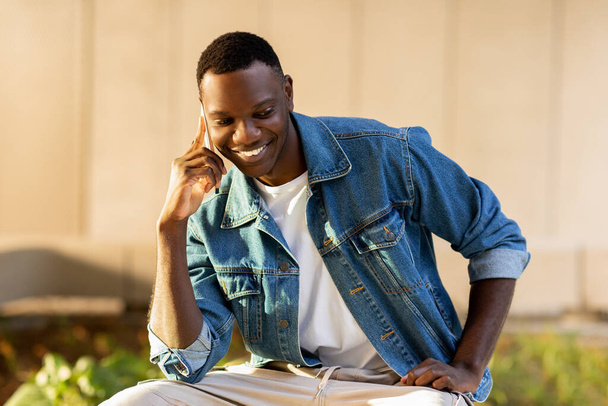 Boldog jóképű, fiatal fekete srác divatos alkalmi ruhában telefonálgat az utcán. Pozitív afro-amerikai diák beszél okostelefonon és mosolyog. Kommunikációs elv - Fotó, kép