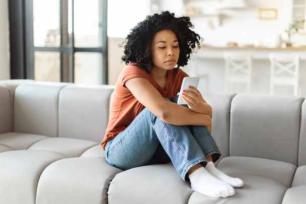 Молода депресивна чорна жінка сидить на дивані і дивлячись на екран смартфона, нескінченно багаторічна афроамериканка чекає на важливий дзвінок або смс, відчуваючи себе самотньо і сумно, тримаючи мобільний телефон - Фото, зображення