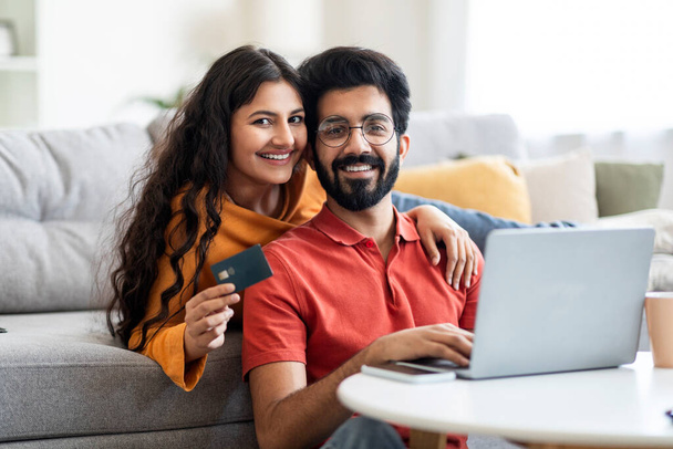 İnternet alışverişleri. Laptop ve kredi kartıyla gülümseyen Hintli çiftin portresi, mutlu genç Doğu eşleri evde birlikte rahatlarken online alışveriş için bilgisayar kullanıyorlar. - Fotoğraf, Görsel