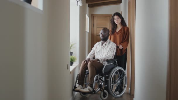 Πλήρης εικόνα μαύρου άνδρα σε αναπηρική καρέκλα με τη βοήθεια νεαρής καυκάσιας γυναίκας στο πλευρό του και οι δύο κοιτάζουν την κάμερα - Πλάνα, βίντεο