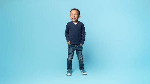 Полнометражный снимок счастливого африканского мальчика-американца, держащегося за руки в карманах, стоящего на синем фоне студии и улыбающегося в камеру, панораму, свободное пространство - Фото, изображение
