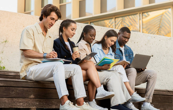 Мультикультурная группа студентов юношей и девушек, сидящих в университетском городке, готовящихся к занятиям, делающих заметки в блокнотах, использующих цифровые планшеты и ноутбуки, пьющих кофе на вынос - Фото, изображение