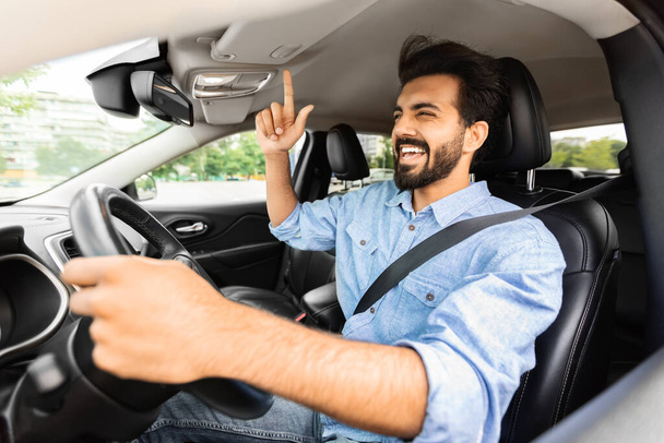 Emozionale millenario bel ragazzo barbuto autista arabo divertirsi durante il viaggio in auto, ascoltare musica e cantare con amore, muovendo la mano, sparato dal finestrino laterale aperto di auto - Foto, immagini