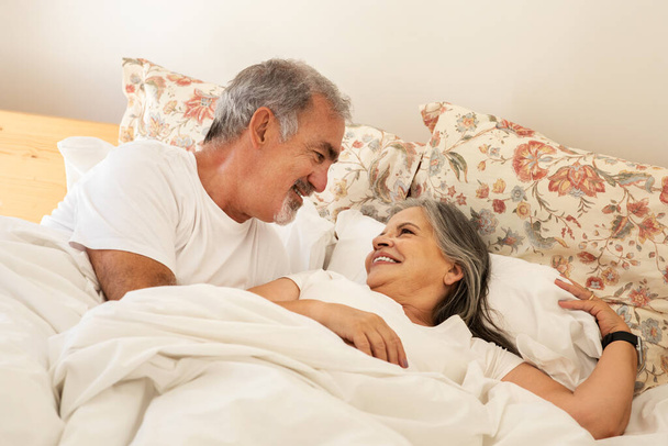 Улыбающийся счастливый старый кавказский муж просыпается с женой в постели, наслаждаясь вместе добрым утром в спальне. Выходные и свободное время, отношения и любовь, образ жизни, отдых, отдых - Фото, изображение