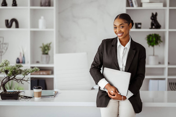Un homme d'affaires prospère. Portrait de femme d'affaires noire heureuse avec ordinateur portable dans les mains debout penché à la table dans le bureau, espace libre. Femme entrepreneur souriant à la caméra - Photo, image
