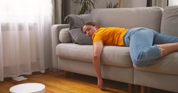 Mujer muy cansada durmiendo en el sofá en casa mientras robot aspirador trabajando en el suelo. Imágenes de alta calidad 4k - Imágenes, Vídeo