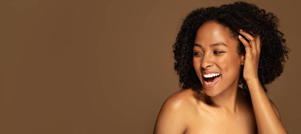Gesichtspflege, Schönheitspflege, ästhetische Medizin, Kosmetologie. Glückliche halbnackte afrikanisch-amerikanische junge Frau, die ihren Kopf berührt und lacht, isoliert auf braunem Hintergrund, Kopierraum, Banner - Foto, Bild