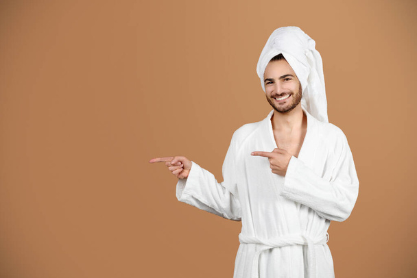 Erkek Güzellik Teklifi. Fotokopi uzayını işaret eden mutlu Latin genç adam, şımartma ve kişisel bakım için kozmetik reklamları yapan, beyaz bornoz giyen ve kahverengi arka planda havluyla gezen - Fotoğraf, Görsel