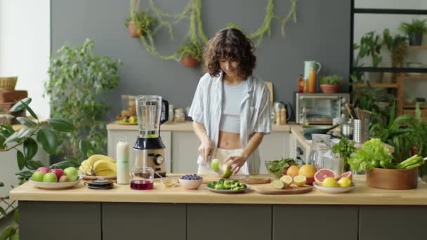 Mittlere Langzeitaufnahme einer jungen Frau, die am Küchentisch frische grüne Äpfel schneidet und zu Hause Frucht-Smoothie zubereitet - Filmmaterial, Video