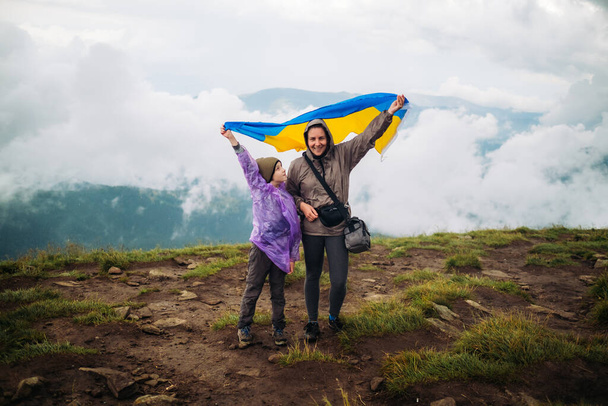 Szczęśliwa kobieta z dzieckiem stoi z narodową ukraińską flagą na szczycie góry Hoverla. Widok na Karpaty w pochmurny dzień. Koncepcja turystyki pieszej, determinacji, samorealizacji - Zdjęcie, obraz