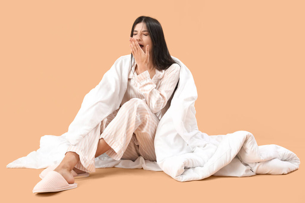 Beautiful young woman with soft blanket yawning on pale orange background - Photo, Image
