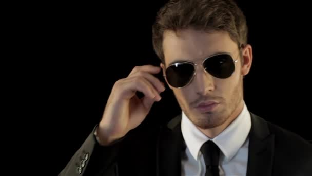 Young caucasian man looking over his sunglasses like special agent. Brunette man in black suite slyghtly lowering his aviator sunglasses on his face. Man taking his sunglasses by the rim isolated - Footage, Video