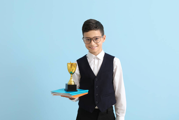Niño de escuela inteligente con taza de premio y copybooks sobre fondo azul claro - Foto, imagen