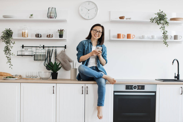 Happy smiling woman in casual wear typing on mobile phone while relaxing in kitchen interior. Joyful asian adult downloading audiobook for entertainment from online library using digital device. - Foto, Bild