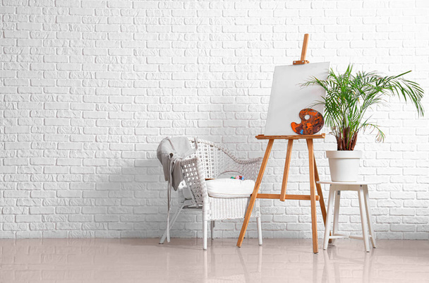 Ξύλινο καβαλέτο με λευκό καμβά κοντά στον τοίχο από λευκό τούβλο - Φωτογραφία, εικόνα