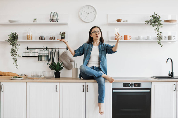 Positive dark haired woman in denim outfit having video call on modern smartphone while sitting on bright kitchen. Asian young lady gesturing and talking during remote communication. - Φωτογραφία, εικόνα
