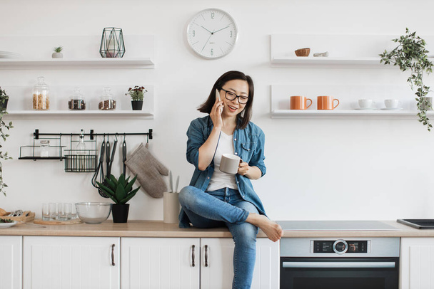 Smiling korean woman with big mug in hand being involved in interesting conversation on mobile on kitchen countertop. Carefree barefoot adult sharing news about birthday plans while having tea. - Photo, image