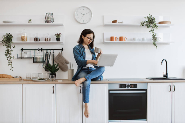 Asian brunette lady with tea cup in hand holding laptop on left knee crossed over right leg in kitchen interior. Relaxed businesswoman enjoying comfort of countertop while teleworking from home. - Photo, image