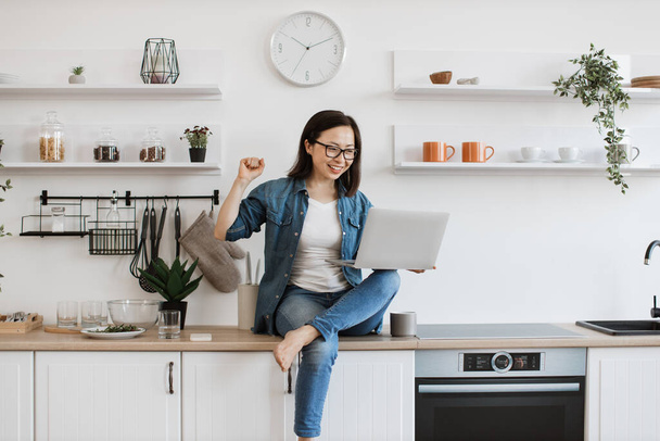 Overjoyed chinese woman in glasses showing victory gesture with raised fist while looking at laptop screen in kitchen. Enthusiastic business lady in denim clothes experiencing joy after big win. - Foto, Imagem